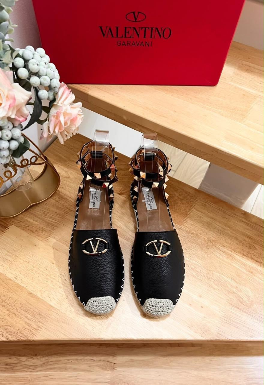 black grained leather Valentino Garavani Rockstud espadrille flats featuring a prominent gold-tone VLogo emblem on the toe and signature pyramid-studded double ankle straps